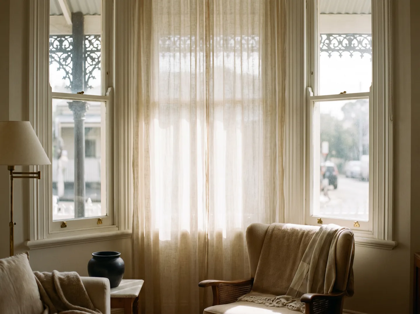 Tall sash windows in a terrace house dressed with a sheer curtain, the street outside softened to silhouettes
